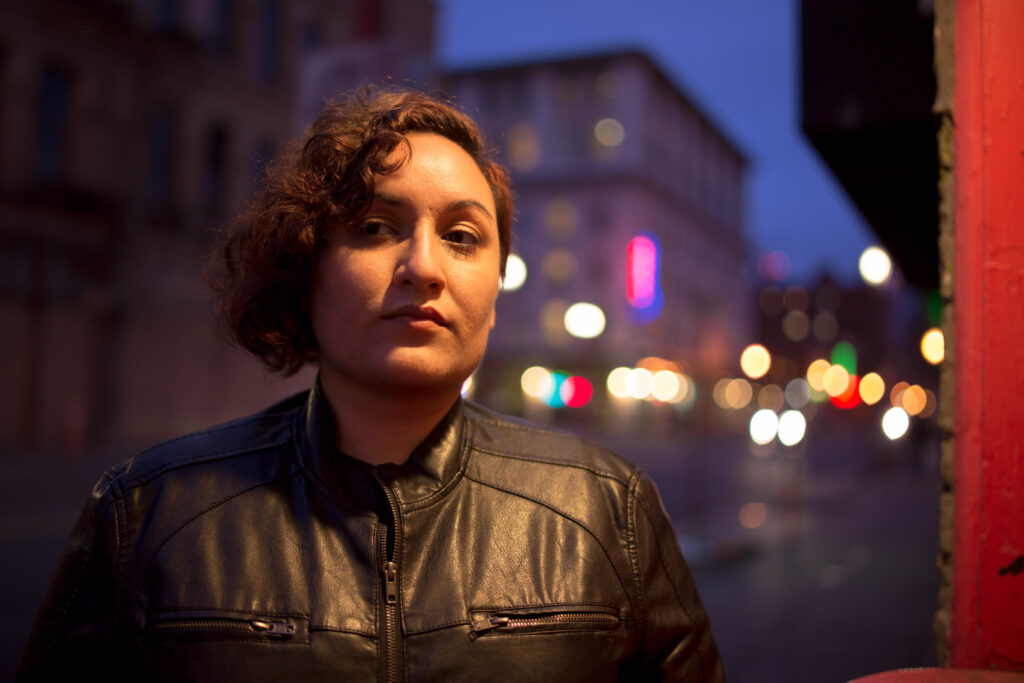 A headshot of Sloane Leong, a cartoonist of mixed indigenous ancestries looking off camera. She has a short, curly haircut and is wearing a black jacket standing in front of a city background that is blurred, and has neon pink, purple and blue behind.