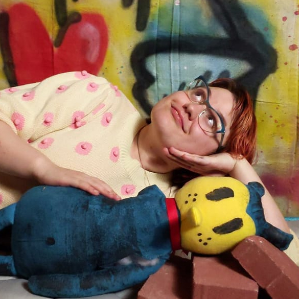 a headshot of Sage Coffey, a trans nonbinary cartoonist holding a plushie. They are looking off screen playfully with a hand against their cheek. Behind them is a wall with spray painted art on it.