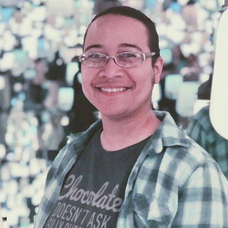 A photo of Jasmine Walls shows her smiling at the camera. She has glasses and short hair and is wearing a shirt about loving chocolate with a flannel over it.