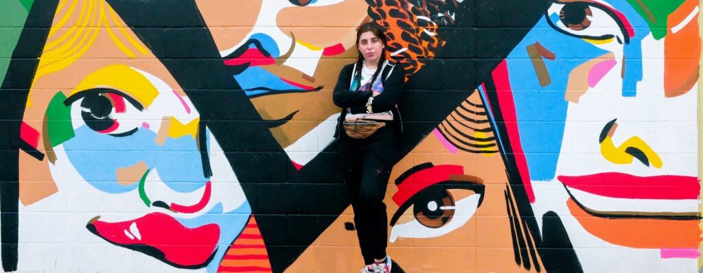 A photo of Molly Mendoza standing in front of a street mural with faces in various colors. Molly has their hair in pigtails, their arms are crossed, and they look extremely cool. Photo credit: Dani Townsend PDX