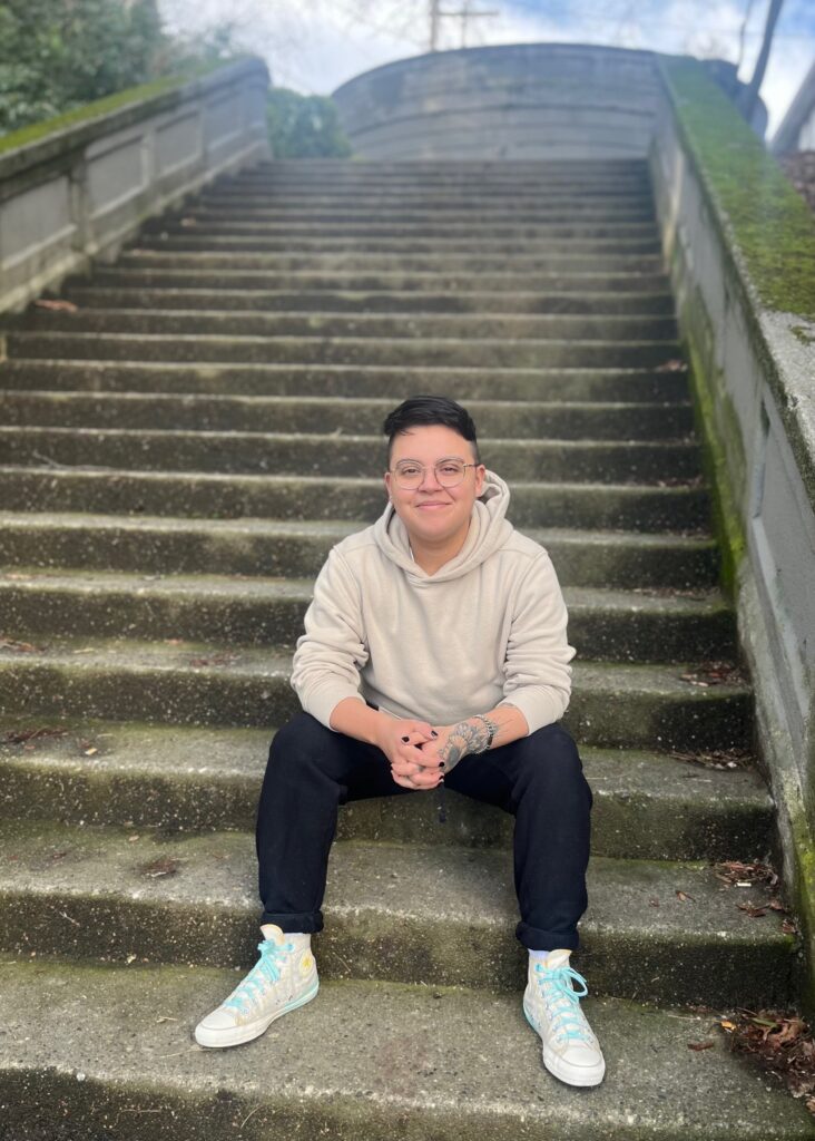 A photo of queer Chicanx comic artist Teo DuVall shows them sitting on a set of concrete stairs, smiling gently in a tan hoodies. They have tattoos on their arm.