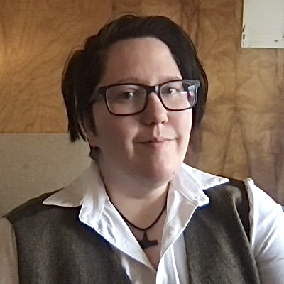 A headshot of author Sarah Edmonds shows them looking at the camera, wearing glasses. They have short dark hair and light skin.