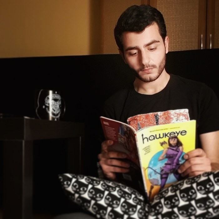 A picture of Anas Abdulhak, a Syrian comic writer reading Hawkeye with a mug to the side. They have a short beard and mustache and are looking down away from the camera.