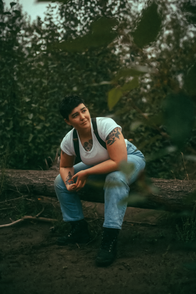 A photo of Ren Strapp, creator of HOW COULD YOU. Ren is a gender nonconforming lesbian with short brown hair and cool arm tattoos. She is looking off camera, surrounded by the woods.